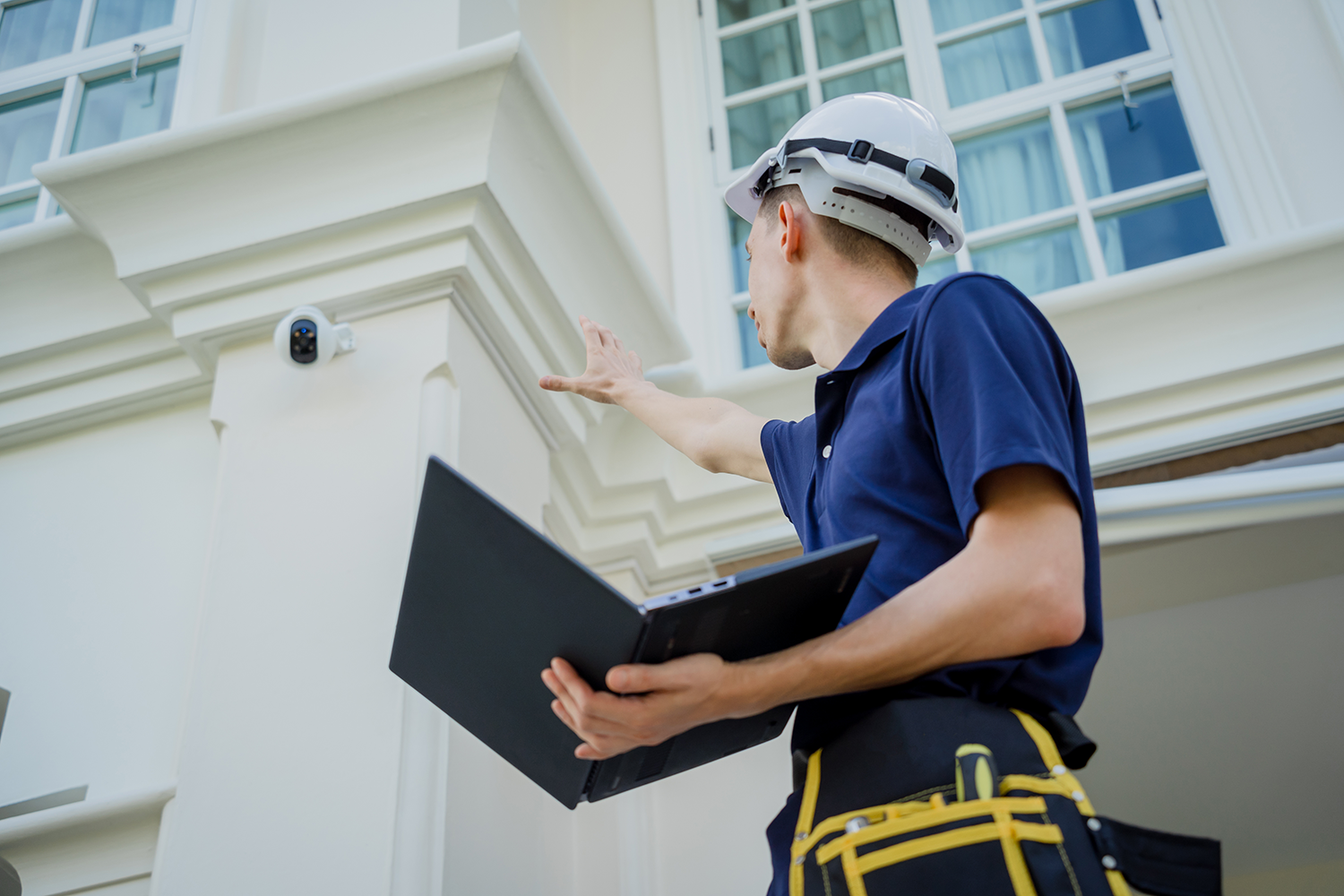 A man in a hardhat holding a computer, gesturing at an exterior security camera