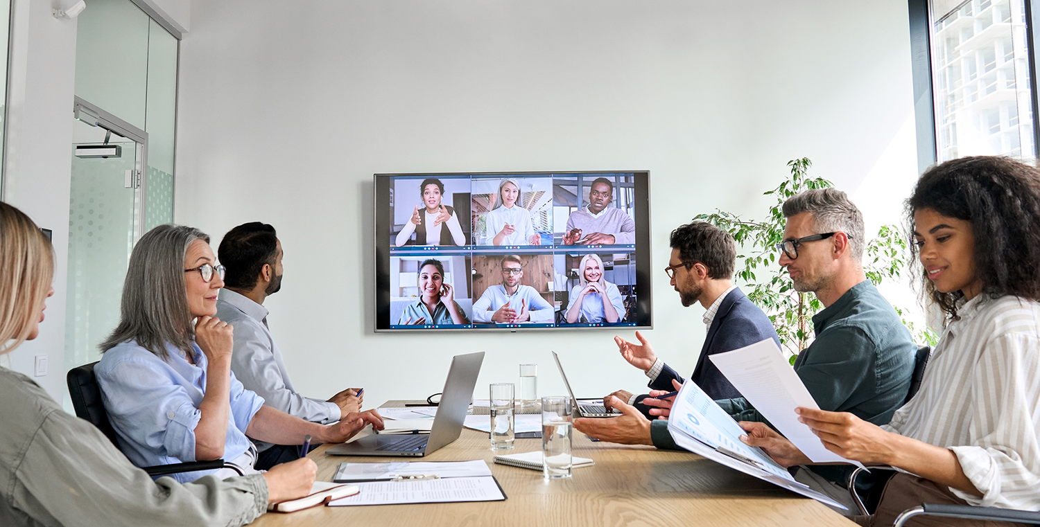 A group of business-people in a meeting room, with more people on a monitor