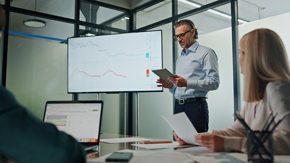 A man leading a meeting in front of a tv showing data graphs